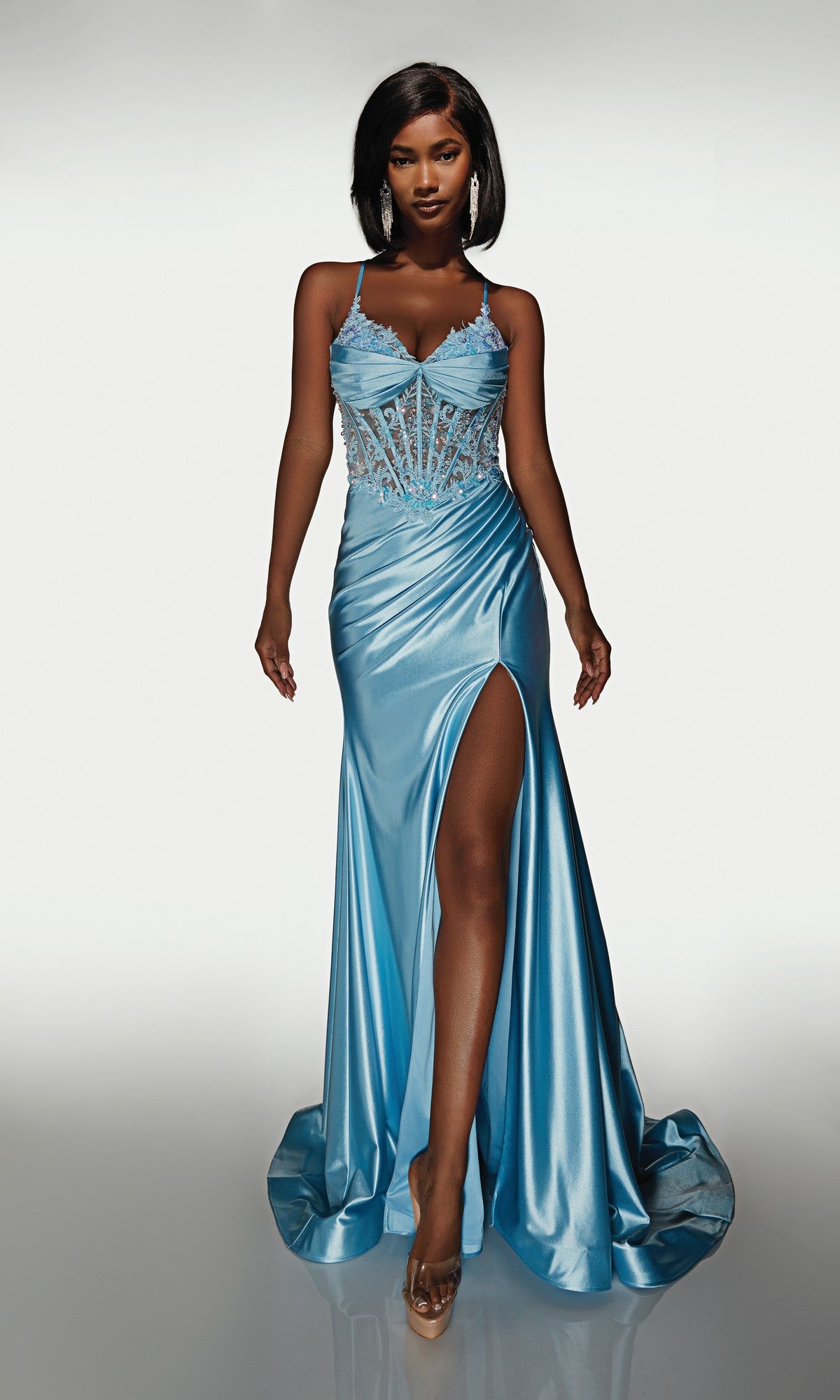 A woman poses in the Alyce 62059 long formal dress, featuring light blue satin, a sheer embellished corset bodice, and a high slit, capturing the modern allure of this standout prom gown against a plain background.